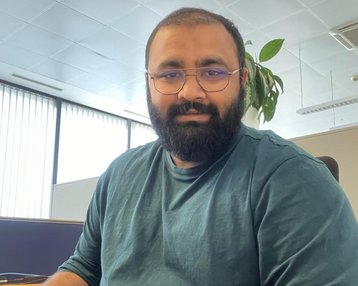 A man with a beard and glasses sits at a desk typing on a keyboard. He is wearing a green top and there is a plant in the background. Daylight streams through large windows.