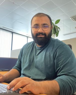 A man with a beard and glasses sits at a desk typing on a keyboard. He is wearing a green top and there is a plant in the background. Daylight streams through large windows.