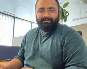 A man with a beard and glasses sits at a desk typing on a keyboard. He is wearing a green top and there is a plant in the background. Daylight streams through large windows.