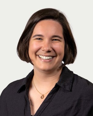 A smiling person with short brown hair is wearing a black shirt and a necklace with a symbol. The background is neutral.