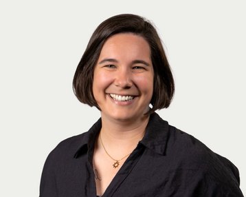 A smiling person with short brown hair is wearing a black shirt and a necklace with a symbol. The background is neutral.
