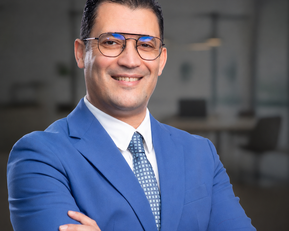 A man in a blue suit with glasses smiles as he stands with his arms crossed in front of a modern office backdrop.