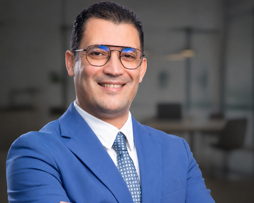 A man in a blue suit with glasses smiles as he stands with his arms crossed in front of a modern office backdrop.