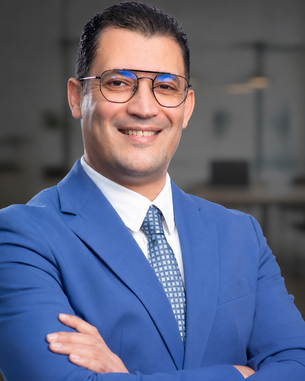 A man in a blue suit with glasses smiles as he stands with his arms crossed in front of a modern office backdrop.