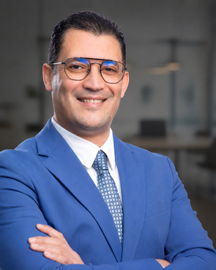 A man in a blue suit with glasses smiles as he stands with his arms crossed in front of a modern office backdrop.