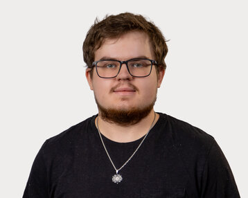 A young man with glasses and a beard is wearing a black T-shirt and a necklace. He is looking directly at the camera.