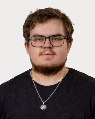 A young man with glasses and a beard is wearing a black T-shirt and a necklace. He is looking directly at the camera.