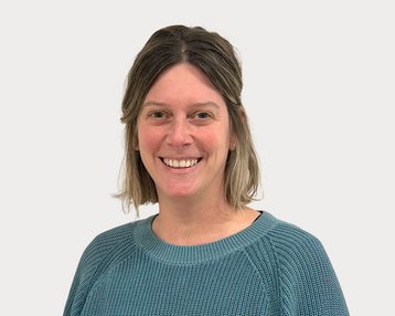 A smiling person with medium-length blonde hair is wearing a blue sweater. The background is neutral and light.