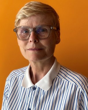 Eine Frau mit kurzen, blonden Haaren und einer gestreiften Bluse steht vor einem orangefarbenen Hintergrund. Sie trägt eine modische Brille.