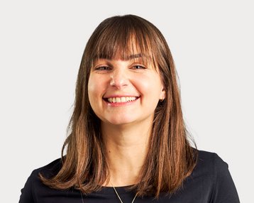 A smiling woman with long brown hair is wearing a black top. The background is neutral and bright.