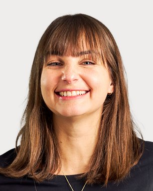 A smiling woman with long brown hair is wearing a black top. The background is neutral and bright.