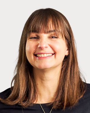 A smiling woman with long brown hair is wearing a black top. The background is neutral and bright.