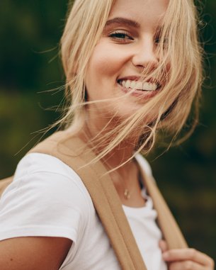Young woman with blonde hair and a smiling face, wearing a white T-shirt and carrying a beige bag. The background features a blurred green.