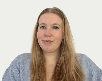 A woman with long blonde hair is wearing a gray sweater and looking directly at the camera. The background is neutral.