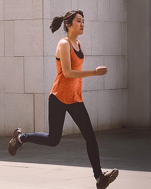 A woman in an orange top and black leggings is energetically running in a modern, minimalist setting.