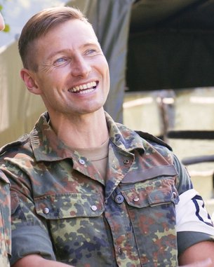 Three soldiers in uniform are standing outside and chatting. They are smiling and appear relaxed, with a tent visible in the background.