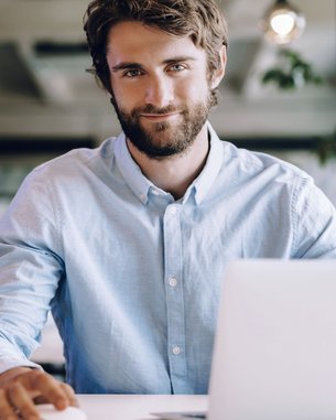 A man with a beard is sitting at a table in a modern office, smiling and working on a laptop. A notebook and a mouse are on the table. Plants are visible in the background.