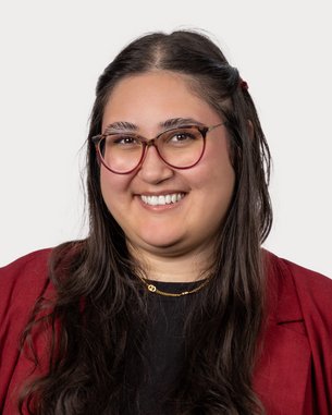 A smiling woman with long, dark hair is wearing a red jacket and a black blouse. She has glasses and is standing in front of a neutral background.