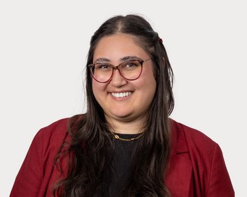 A smiling woman with long, dark hair is wearing a red jacket and a black blouse. She has glasses and is standing in front of a neutral background.