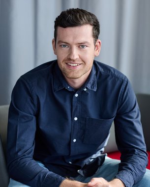 A young man in a dark shirt sits on a chair, smiling with his hands resting on his knees.