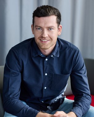 A young man in a dark shirt sits on a chair, smiling with his hands resting on his knees.