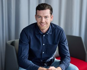 A young man in a dark shirt sits on a chair, smiling with his hands resting on his knees.