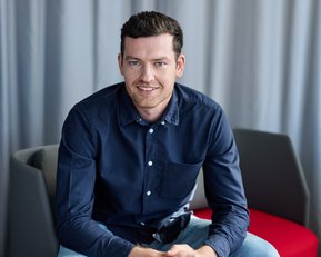 A young man in a dark shirt sits on a chair, smiling with his hands resting on his knees.