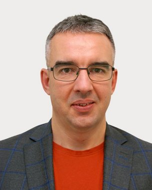 Man with glasses, gray hair, and a gray checkered blazer, wearing a red T-shirt. Neutral background.