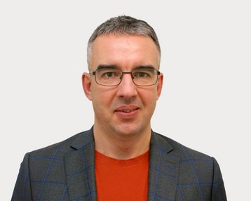 Man with glasses, gray hair, and a gray checkered blazer, wearing a red T-shirt. Neutral background.