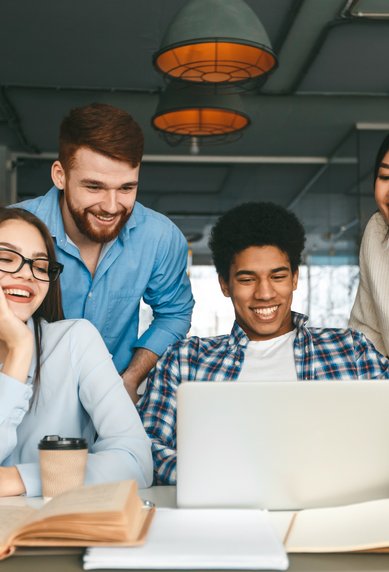 Six young adults are sitting at a table, looking at a laptop. They seem engaged and are laughing as they work together on a project. Books and writing supplies are scattered across the table.