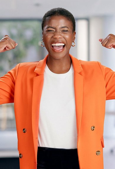 Eine Frau in einem orangefarbenen Blazer lächelt und hebt ihre Arme in einer starken Pose. Der Hintergrund zeigt einen modernen Büroraum mit Pflanzen und Möbeln.