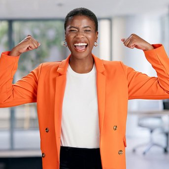 Eine Frau in einem orangefarbenen Blazer lächelt und hebt ihre Arme in einer starken Pose. Der Hintergrund zeigt einen modernen Büroraum mit Pflanzen und Möbeln.
