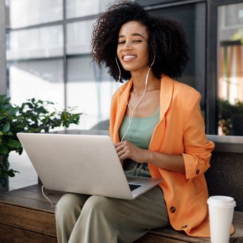 A woman with curly hair is sitting at an outdoor table, wearing an orange blazer and listening to music while working on her laptop.