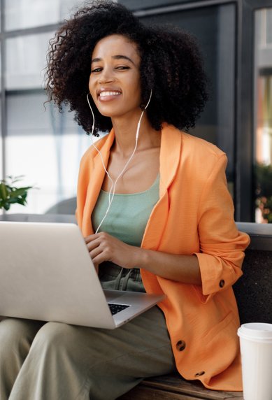 A woman with curly hair is sitting at an outdoor table, wearing an orange blazer and listening to music while working on her laptop.