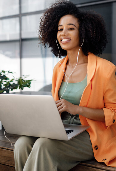 A woman with curly hair is sitting at an outdoor table, wearing an orange blazer and listening to music while working on her laptop.