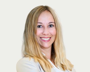 A smiling woman with long blonde hair is wearing a light blazer and standing in front of a neutral background.