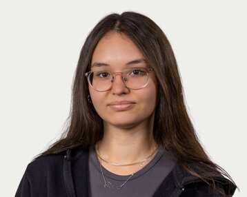 Young woman with long, straight hair and glasses, wearing a black hoodie and a gray blouse. She is looking directly at the camera.
