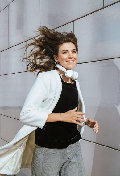 A woman with headphones walks along a wall, wearing a white blazer and has long, flowing hair.