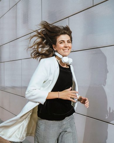 A woman with headphones walks along a wall, wearing a white blazer and has long, flowing hair.