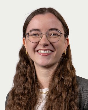 A smiling woman with long, curly hair and glasses is wearing a blazer. The background is neutral and bright.