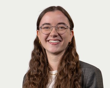 A smiling woman with long, curly hair and glasses is wearing a blazer. The background is neutral and bright.