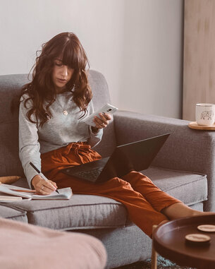 A woman is sitting on a sofa, using a laptop and holding a smartphone in her hand. Next to her, there are documents and a cup on a table. In the background, bookshelves are visible.