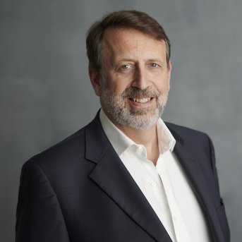 A man with a beard is wearing a dark suit and a white shirt. He stands against a neutral background and has a slight smile.