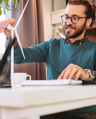 A man is sitting at a desk, working on a laptop while holding a model of a wind turbine in his hand. In the background, plants and a coffee cup are visible.