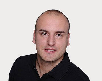 A man with short, smooth hair is wearing a black polo shirt and looking directly at the camera. The background is neutral.