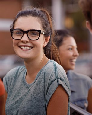 A young woman with glasses smiles at the camera, surrounded by other young people. She is wearing a gray T-shirt and has long, curly hair.