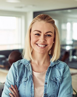 A smiling woman with blonde hair stands in the foreground of a modern office, while in the background, two people are working at a table.