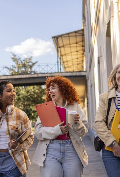 Three women are walking smilingly on a sidewalk, carrying binders and a coffee cup, surrounded by buildings and daylight.