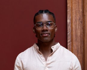 A young man with short, braided hair and glasses is wearing a light-colored shirt and standing in front of a red wall.