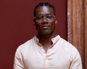 A young man with short, braided hair and glasses is wearing a light-colored shirt and standing in front of a red wall.
